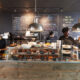 Baristas preparing drinks behind a rustic coffee shop counter with a display of pastries and chalkboard menus, showcasing a cozy, Instagrammable café atmosphere in Destin, Florida.
